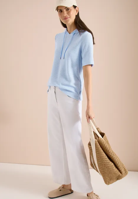 Person in hellblauem Kurzarm-Hoodie, weißer weiter Hose, Basttasche und Slipper vor neutralem Hintergrund.