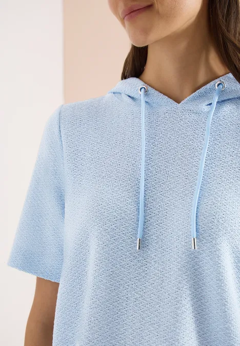 Frau trägt hellblauen kurzärmligen Kapuzenpullover mit strukturierter Oberfläche, Gesicht und Oberkörper teils sichtbar.