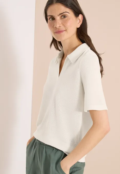 Effen polo met korte mouwen macadamia beige Vrouw in een witte polo met korte mouwen en een groene broek, zijwaarts staand met handen in de zakken.