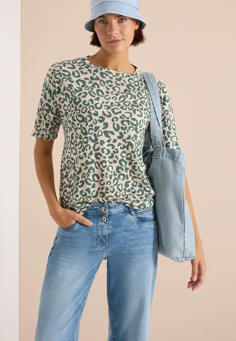 Une personne porte un t-shirt vert à motif léopard, un jean bleu, un bob et un sac en tissu bleu clair.