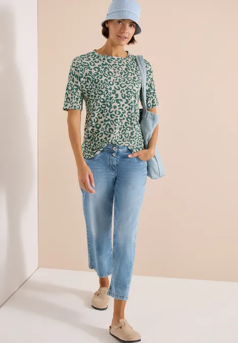 Femme en T-shirt vert à fleurs, jean clair et espadrilles compensées, accessoirisée d'un chapeau et d'un sac bleu pâle.
