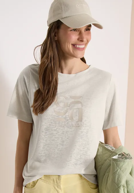 Vrouw met licht T-shirt en pet, houdt een groene jas vast voor een neutrale achtergrond.