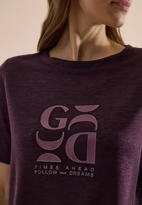 Nahaufnahme eines lila T-Shirts mit stylisiertem 'GA'-Schriftzug und 'Times Ahead Follow Your Dreams'.