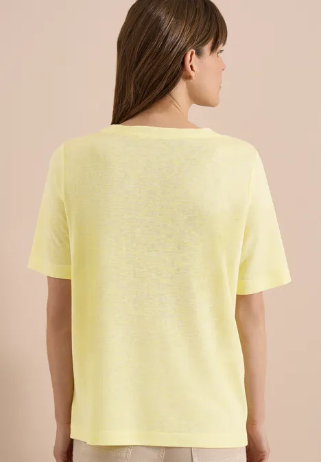 Vrouw van achter gefotografeerd met geel t-shirt en paardenstaart voor een beige achtergrond.