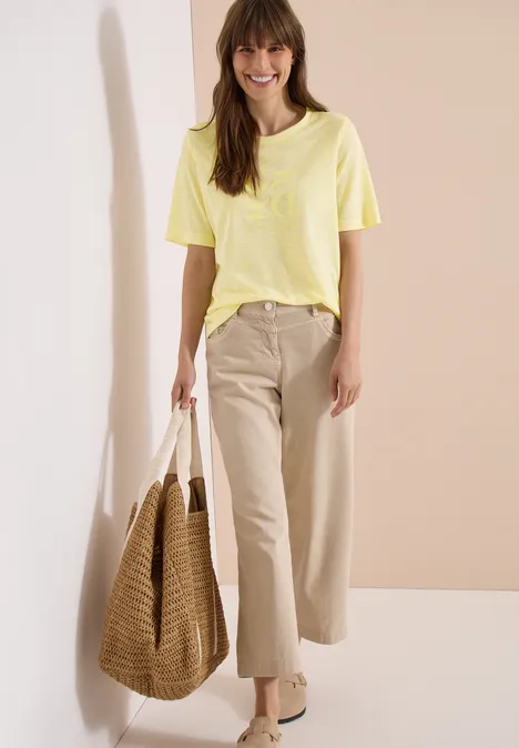 Vrouw in geel T-shirt, beige broek en grote rieten tas voor neutrale achtergrond.