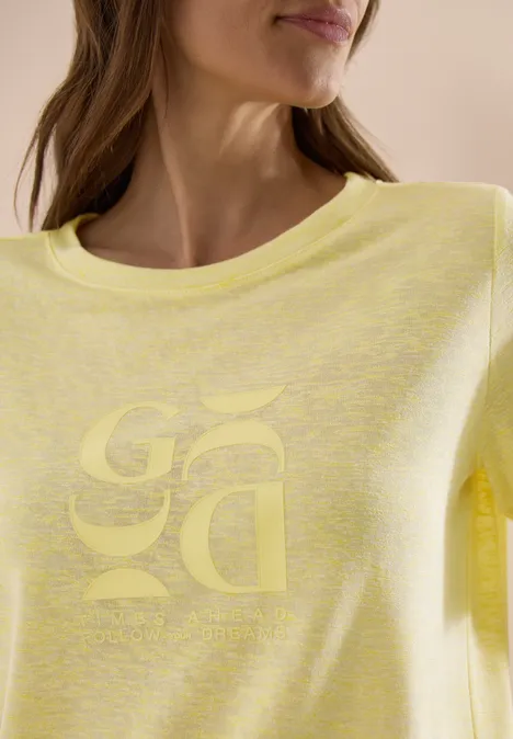 Vrouw in lichtgeel T-shirt met ronde hals en subtiel spiegelend logo op de borst.