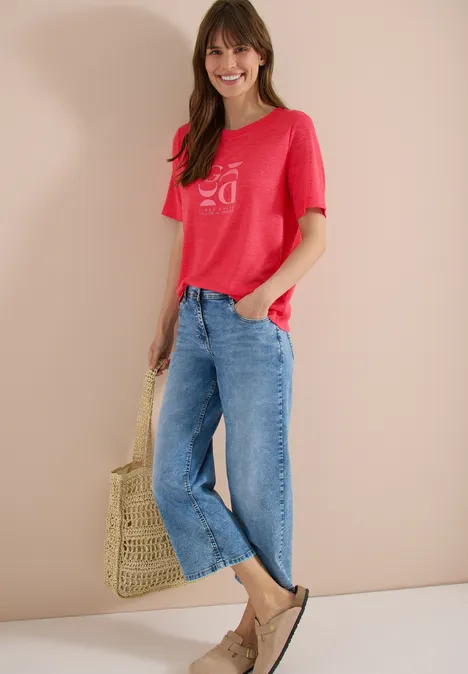 Frau in rotem T-Shirt, weiten blauen Jeans, Strohtasche und Sandalen vor neutralem Hintergrund.