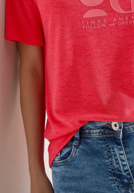 Person trägt rotes T-Shirt mit hellem Print und blaue Jeans, lässiges Outfit