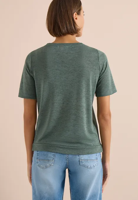 Frau steht mit dem Rücken zur Kamera und trägt ein grünes T-Shirt sowie blaue Jeans.