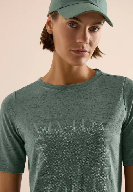 Person trägt grünes T-Shirt und Cap vor hellem Hintergrund, Gesicht unkenntlich.