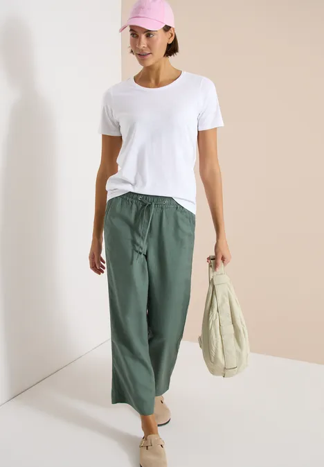 Model draagt wit T-shirt, wijde olijfgroene broek, comfortabele schoenen en houdt een beige tas voor een lichte achtergrond.