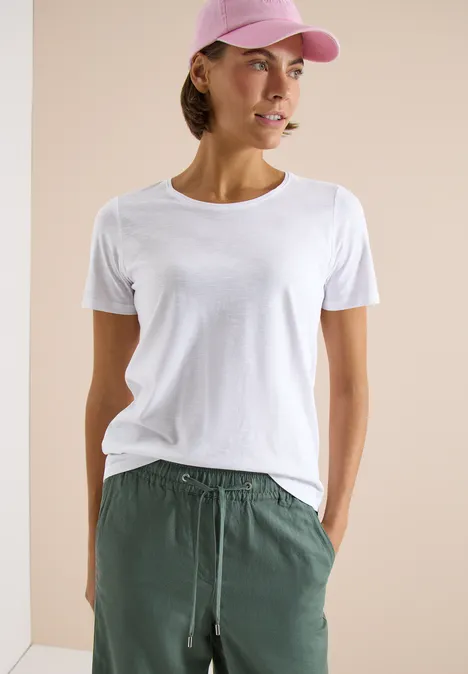 Persoon draagt wit t-shirt, groene broek en roze pet, staand voor een lichte achtergrond.