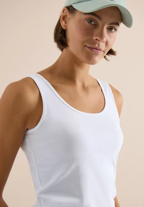 Persoon met een wit mouwloos hemd en baseballpet tegen een neutrale, lichte achtergrond.