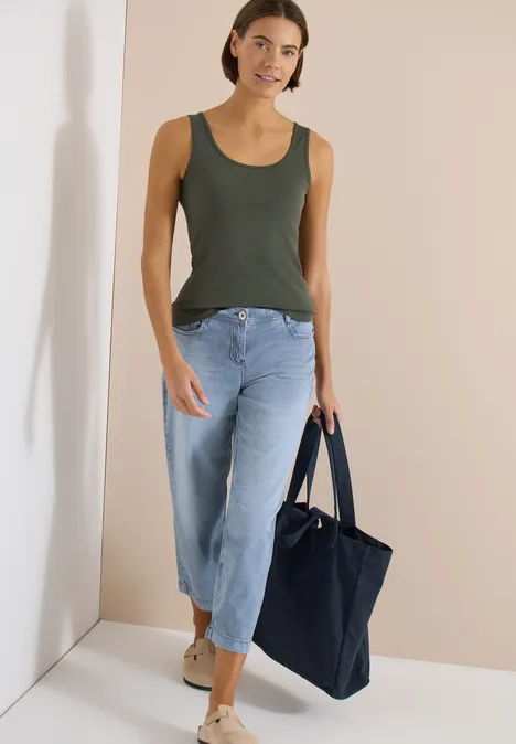 Vrouw in een olijfgroene tanktop, wijde jeans en met een donkerblauwe tas voor een lichte achtergrond.