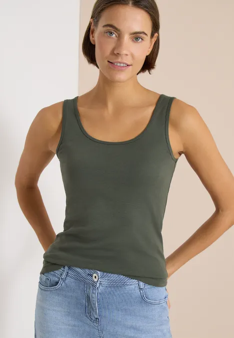 Een vrouw draagt een olijfgroene tanktop en een lichte spijkerbroek tegen neutrale achtergrond.