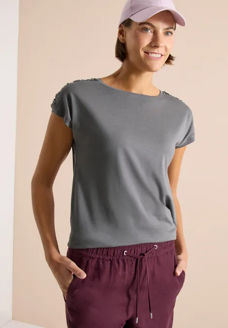 Shirt met korte mouwen en rimpels graphite light grey Vrouw in grijs T-shirt, bordeauxrode joggingbroek en roze pet voor een neutrale achtergrond.