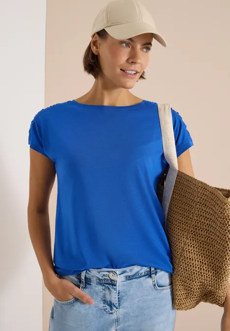 Person trägt blaues T-Shirt, Jeans, weiße Kappe und Strohtasche über der Schulter.
