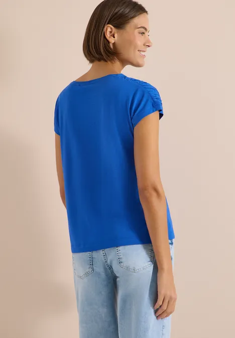 Frau von hinten in blauem T-Shirt und hellen Jeans vor schlichtem, beigem Hintergrund.