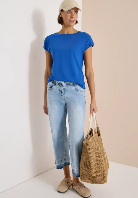 Frau steht in blauem T-Shirt, hellen Jeans, Sneakern und trägt eine große geflochtene Tasche.