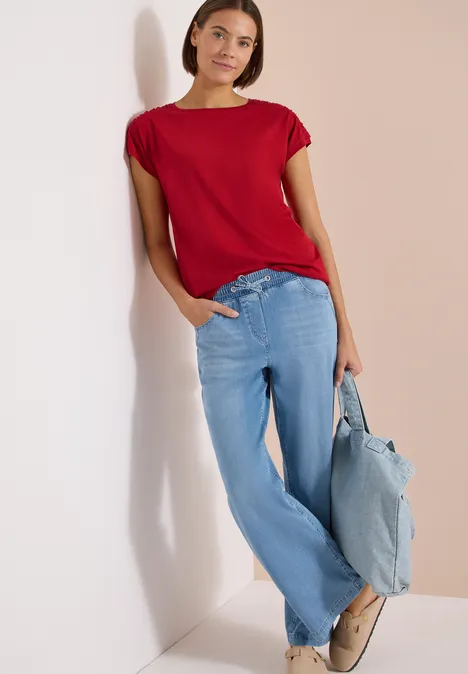 Frau lehnt an weißer Wand, trägt rotes Shirt, weite hellblaue Jeans und hellblaue Tasche.