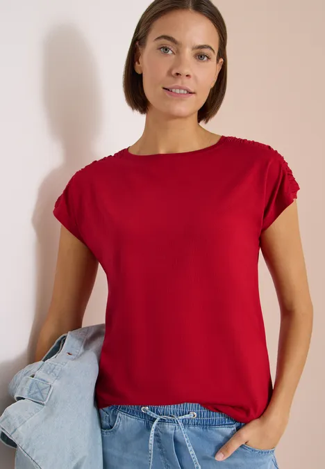 Eine Frau in rotem T-Shirt hält eine blaue Jeansjacke vor hellem Hintergrund.