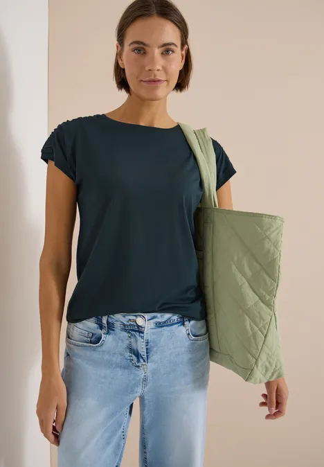 Frau in dunkelblauem T-Shirt, hellen Jeans und grüner Stepp-Umhängetasche.