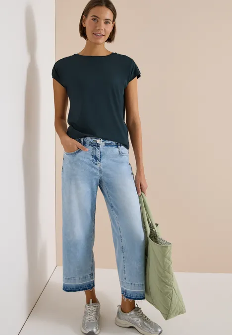 Person trägt schwarzes T-Shirt, weite helle Jeans und hält eine große grüne Stofftasche.