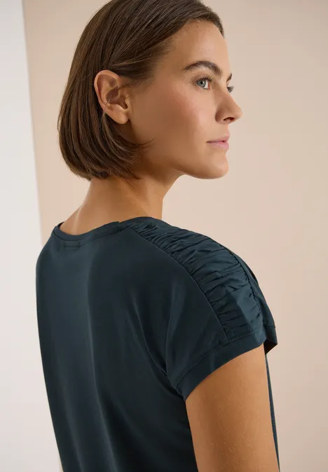 Frau im Profil mit mittellangem Bob und dunkelblauem, gerafftem T-Shirt-Ärmel vor neutralem Hintergrund.