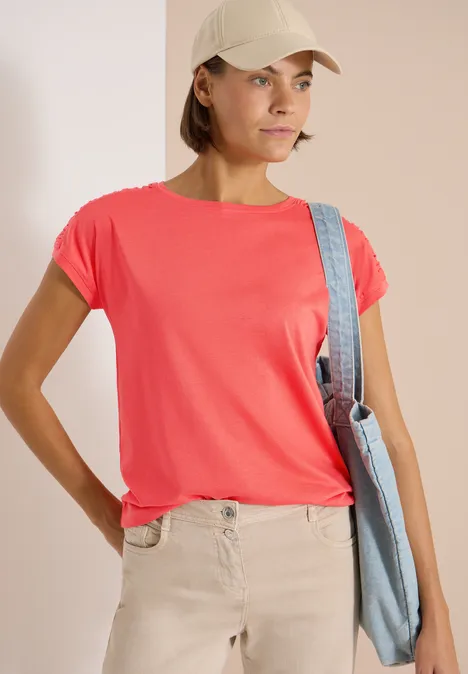Frau in rotem T-Shirt, beiger Hose und Cap, trägt eine Umhängetasche.