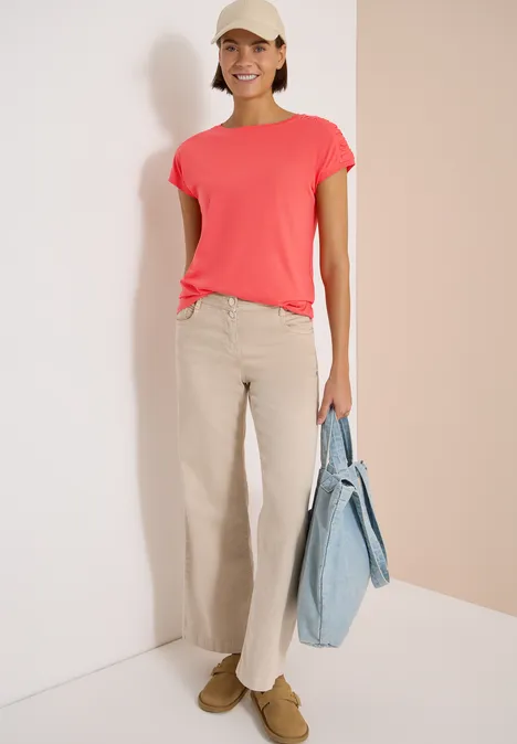Person trägt korallenrotes T-Shirt, beige Hose und hellblaue Tasche vor neutralem Hintergrund.