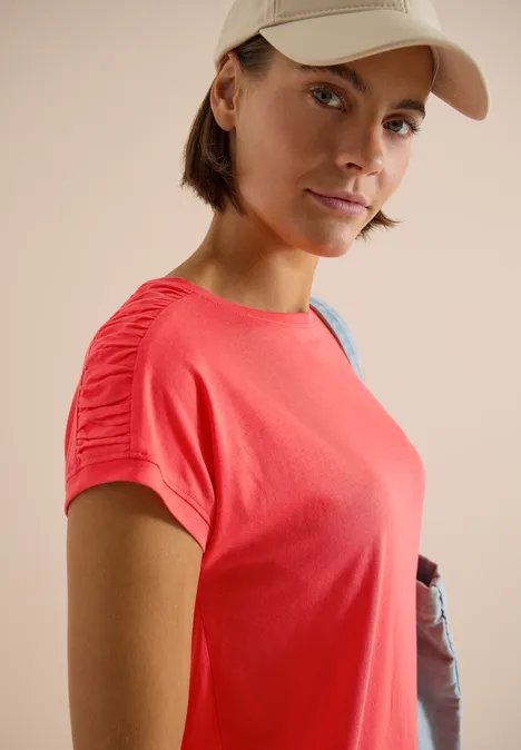 Person in rotem T-Shirt und Baseballcap mit Umhängetasche vor neutralem Hintergrund.