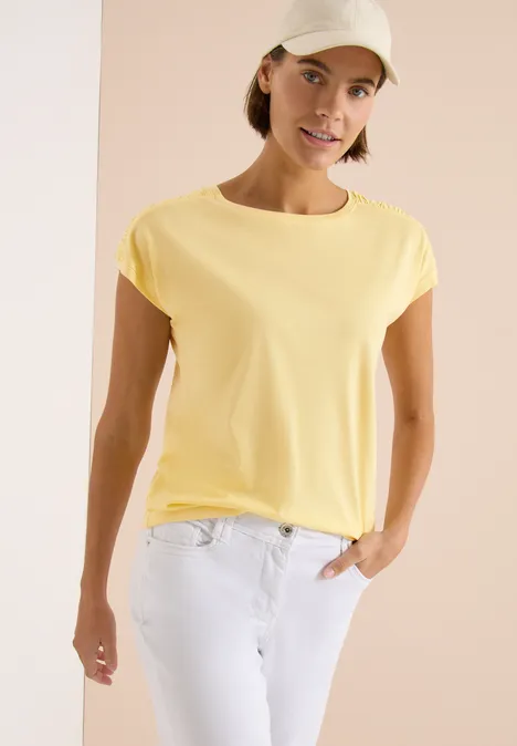 Shirt met korte mouwen en rimpels buttermilk yellow Een vrouw draagt een geel T-shirt, witte jeans en een beige pet, tegen een lichte achtergrond.