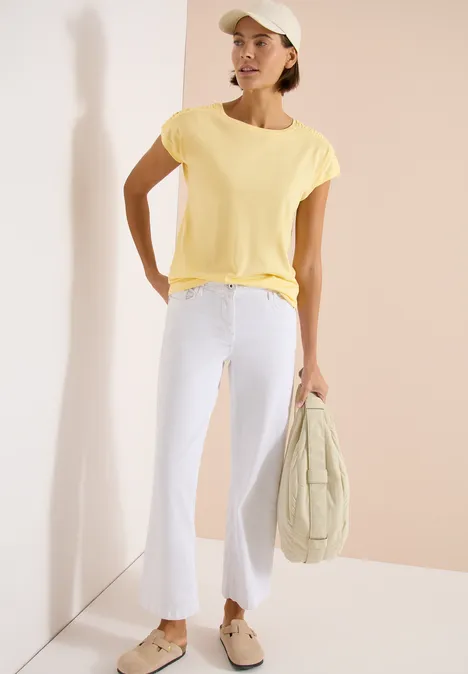 Shirt met korte mouwen en rimpels buttermilk yellow Persoon met geel T-shirt, witte flared broek en beige platformschoenen, houdt gestikte tas vast tegen lichte achtergrond.