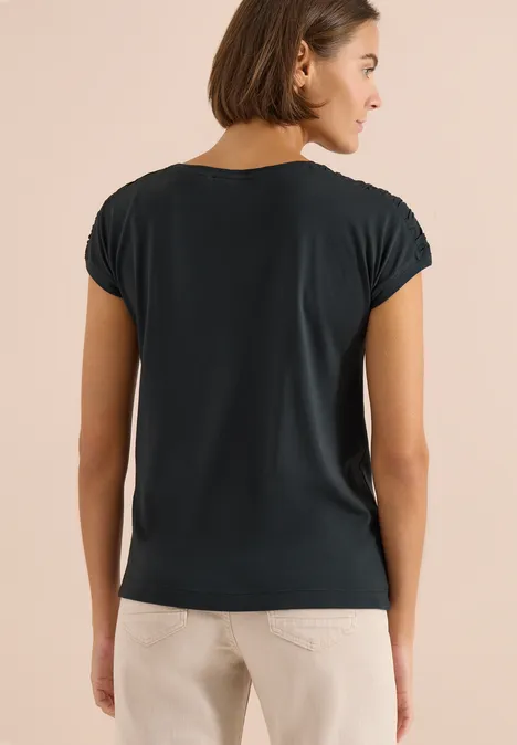 Vrouw van achteren in een donkerblauw t-shirt en lichte broek tegen een neutrale achtergrond.