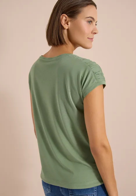 Femme vue de dos portant un t-shirt vert à épaules froncées et un jean devant un fond beige.