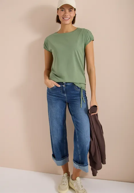 Femme en t-shirt vert, jean bleu retroussé, baskets blanches, tient une veste marron, fond neutre.