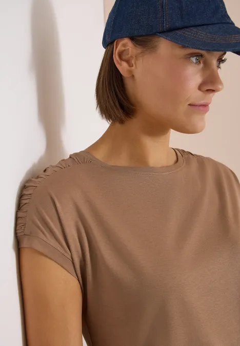 Une personne porte un t-shirt marron à manches courtes froncées et une casquette bleu marine.