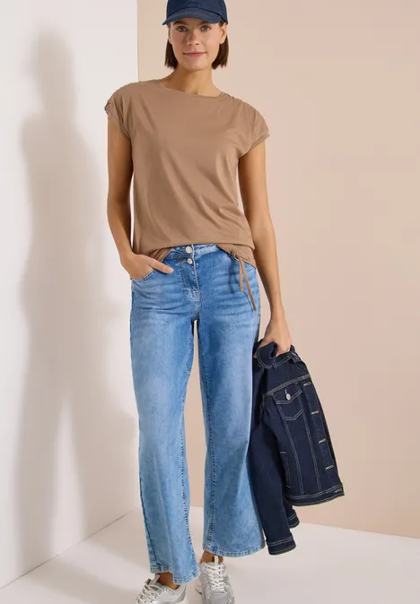 Une femme porte un T-shirt beige, un jean bleu et tient une veste en jean à la main.