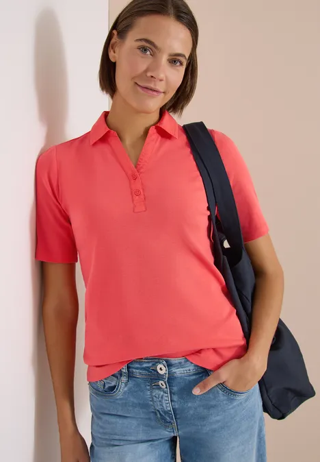 Basic Poloshirt mit Kurzarm shiny coral Person in rosa Poloshirt, Jeans und Schultertasche vor hellem Hintergrund.