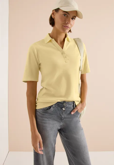 Basis polo met korte mouwen buttermilk yellow Persoon draagt lichtgeel poloshirt, grijze broek en schoudertas tegen neutrale achtergrond.
