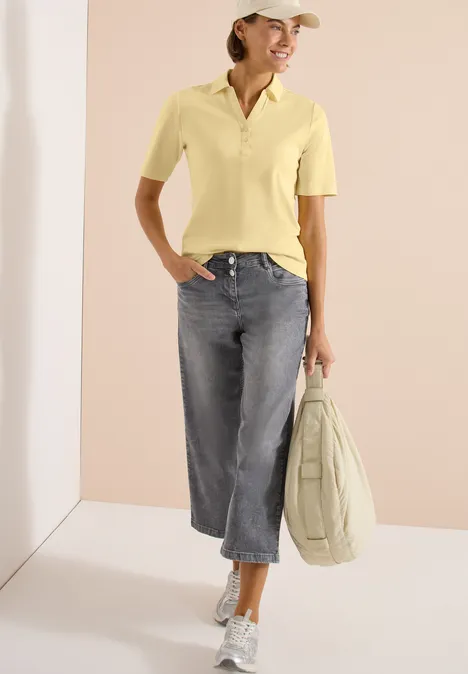 Basis polo met korte mouwen buttermilk yellow Vrouw in gele polo, grijze wijdvallende jeans, witte sneakers, pet en lichte tas in een minimalistisch interieur.