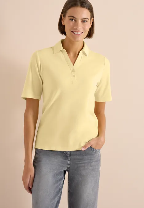 Basis polo met korte mouwen buttermilk yellow Vrouw in geel poloshirt en spijkerbroek, hand in zak, staand voor een lichte achtergrond.