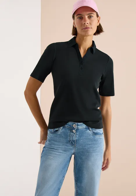 Basis polo met korte mouwen palm breeze green Vrouw in een zwart poloshirt en blauwe jeans, staand voor een lichte achtergrond.