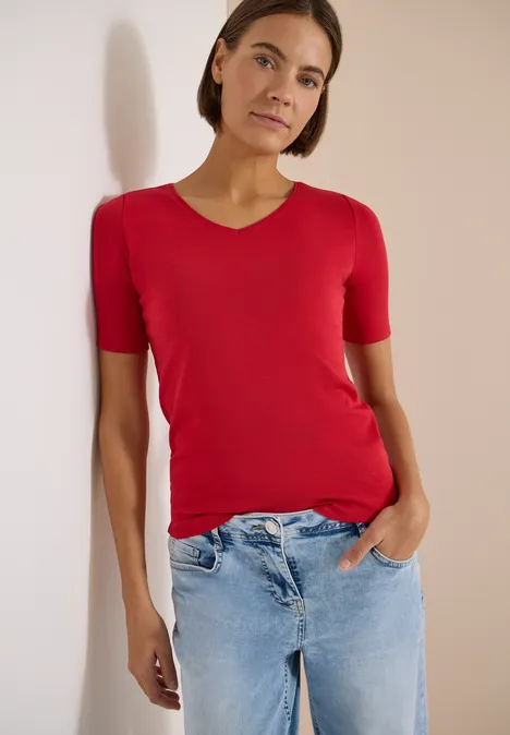 Basic T-Shirt aus Baumwolle tamarillo red Eine Frau in rotem T-Shirt und blauen Jeans steht entspannt mit einer Hand in der Tasche vor hellem Hintergrund.