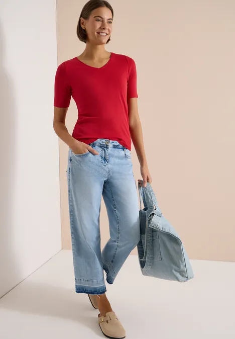 Basic T-Shirt aus Baumwolle tamarillo red Frau trägt rotes Kurzarmshirt, weite hellblaue Jeans, hält lässig eine Jeansjacke, beige Schuhe.