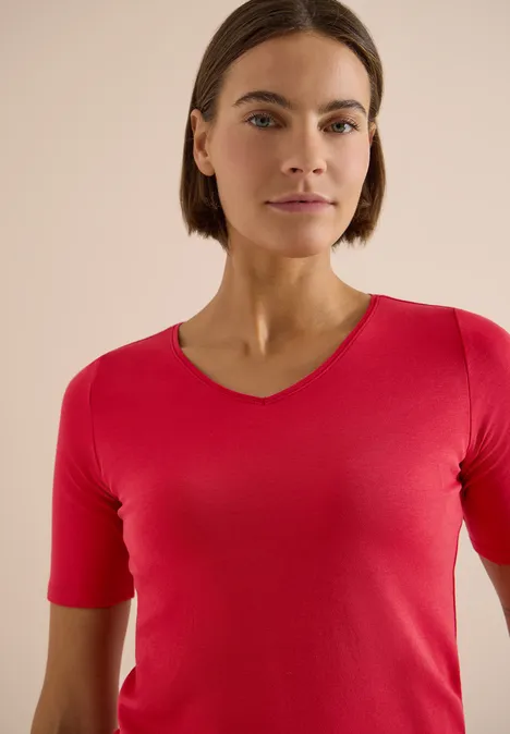 Basic T-Shirt aus Baumwolle tamarillo red Frau mit rotem V-Ausschnittshirt und verschwommenem Gesicht vor beigem Hintergrund.