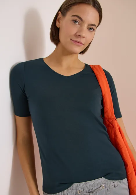 Basic T-Shirt aus Baumwolle deep petrol blue Jemand lehnt entspannt in einem dunkelblauen T-Shirt an einer Wand, mit einer orangenen Tasche über der Schulter.