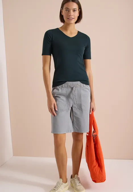 Basic T-Shirt aus Baumwolle deep petrol blue Eine Person trägt ein dunkelgrünes T-Shirt, graue Bermudashorts und hält eine orangefarbene Tasche.