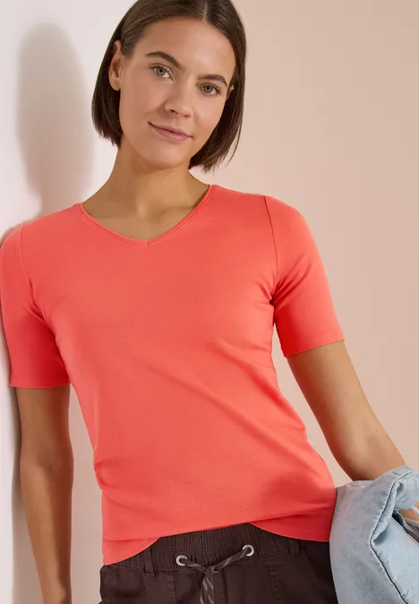 Model draagt een koraalroze T-shirt en houdt een lichtblauwe spijkerbroek vast, neutrale achtergrond.