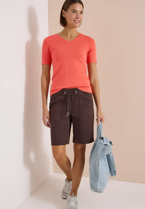 Vrouw in koraalkleurig V-shirt, donkere bermudashort met koord, sneakers en spijkerjas.